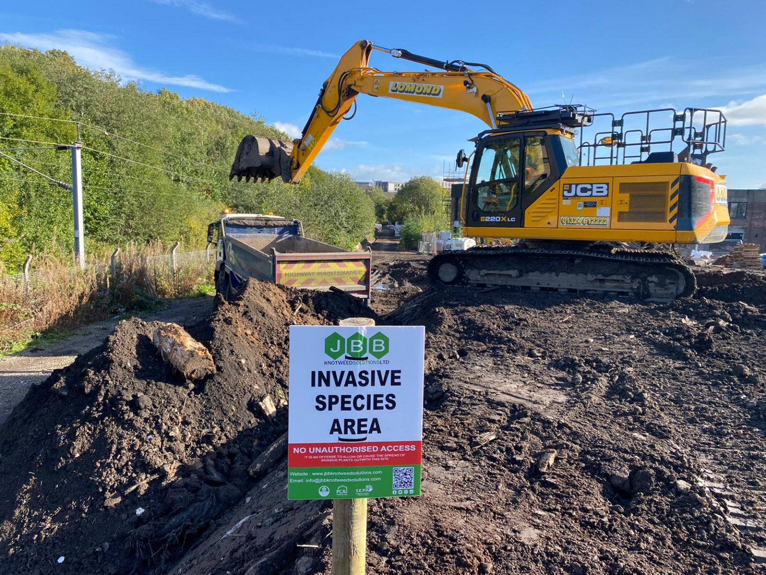 Yellow excavator carrying out work in a JBB Knotweed solutions branded and signposted 'Invasive Species Area'