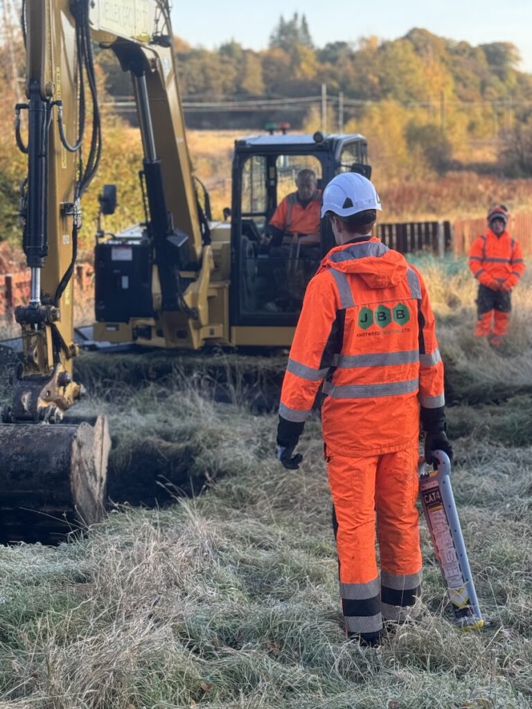 JBB team member with a CAT scan tool to check underground services on a commercial site with a JCB carrying out reinstatement in the background.