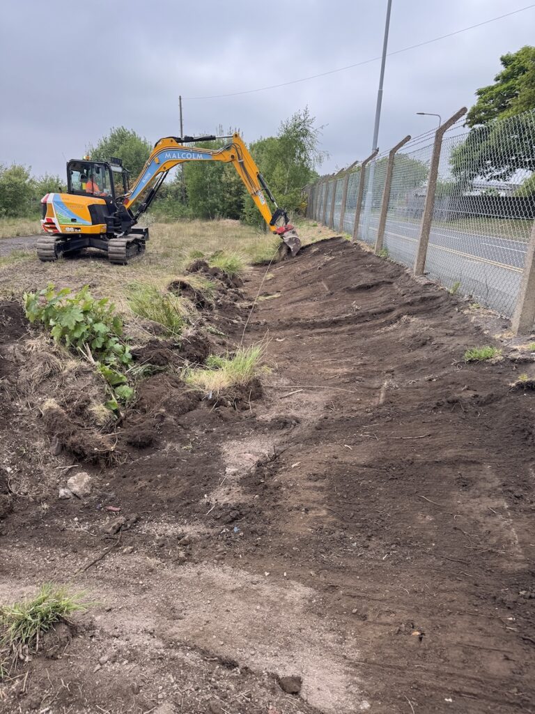 JCB excavator reinstating ground after invasive weed treatment beside metal wire fencing at a commercial site.