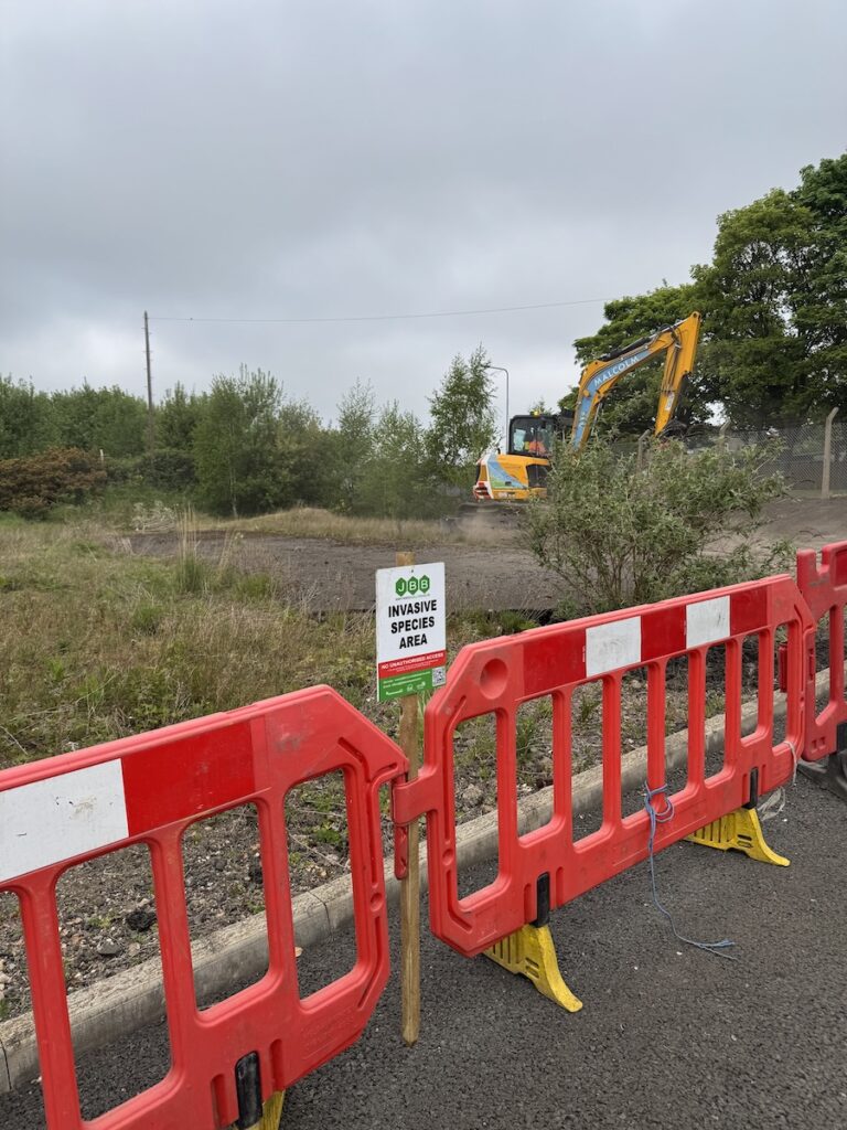 JCB carrying out land reinstatement at a commercial roadside site with JBB invasive weed signage and safety barricades in place.