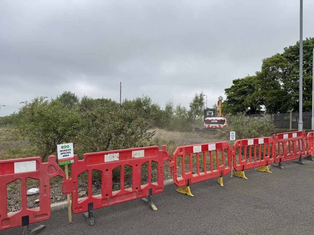 JCB reinstating land at a commercial roadside site with JBB invasive weed signage and safety barricades in place.